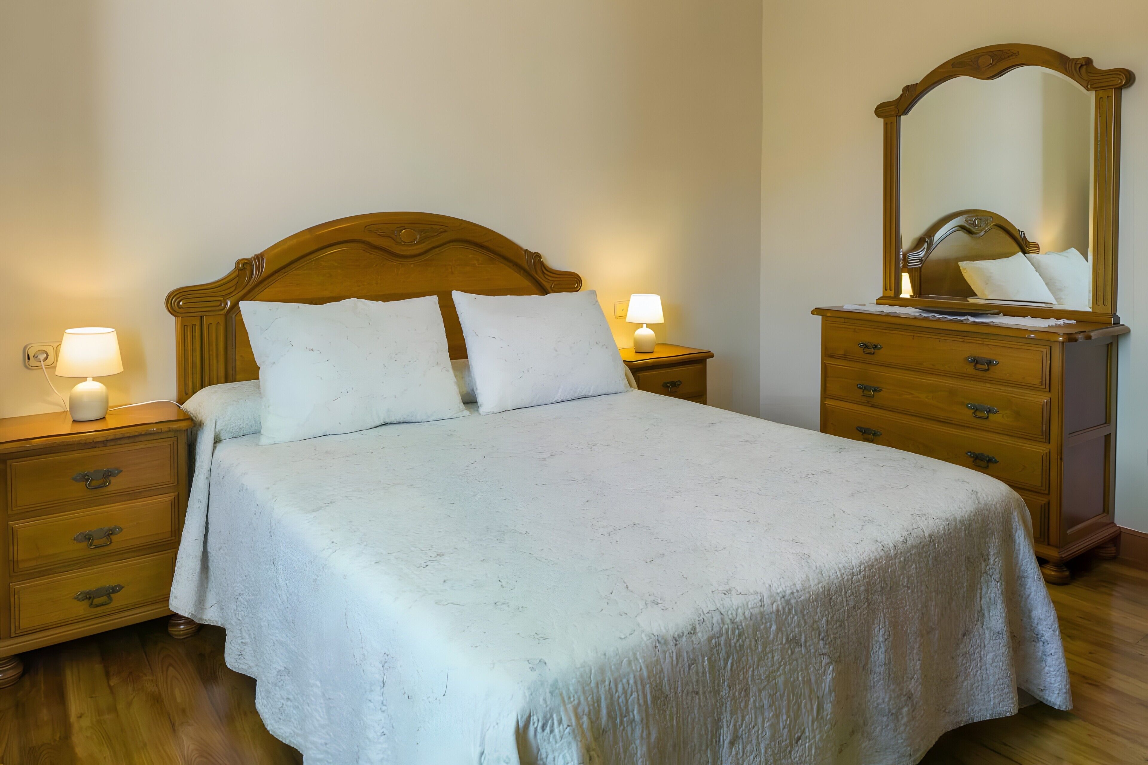 Un letto ben rifatto con una testiera in legno, due lampade da comodino, un comodino in legno con cassetti e uno specchio sulla parete.