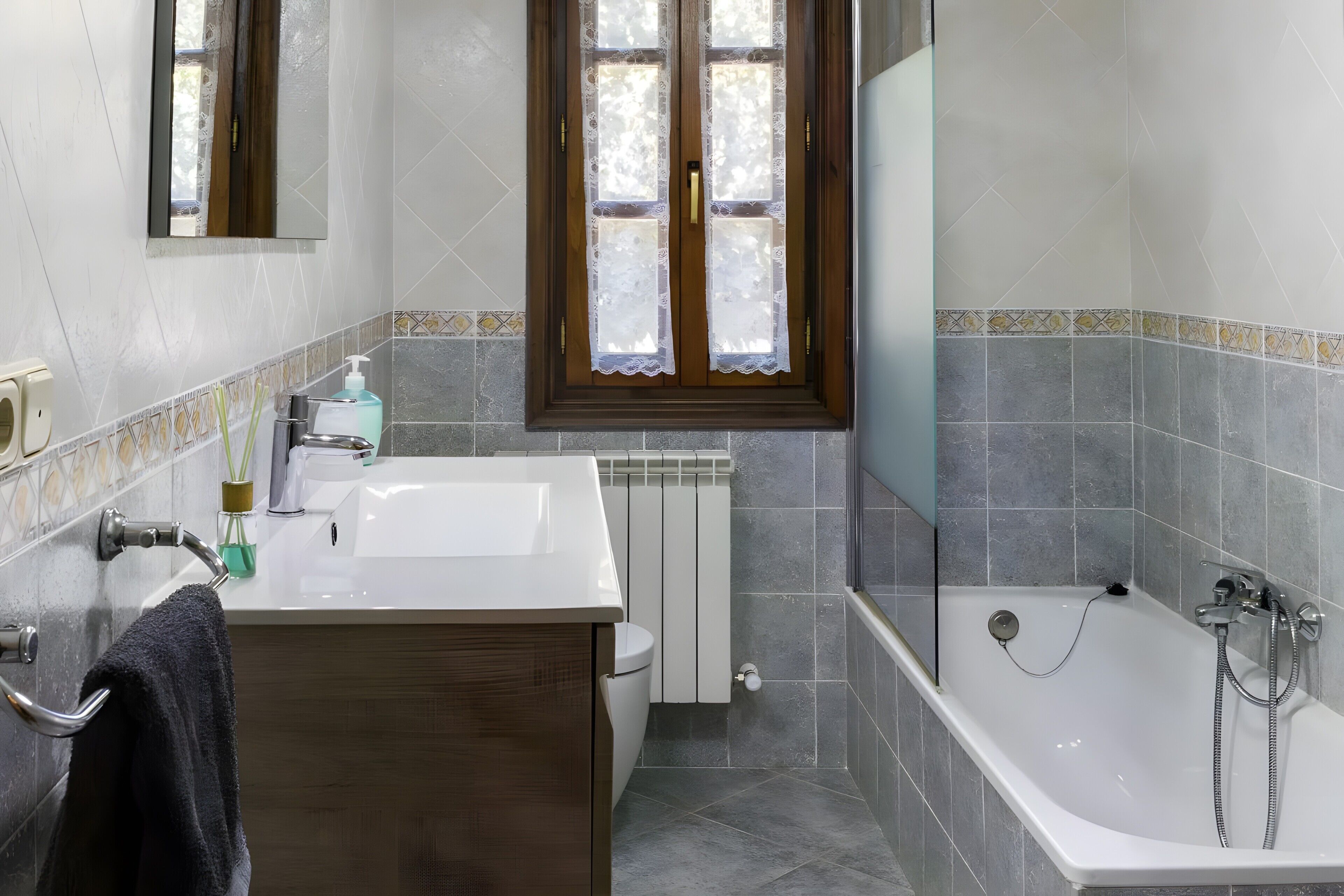 Un baño con lavamanos blanco, tocador de madera, inodoro, bañera con ducha y ventana con cortinas de encaje.