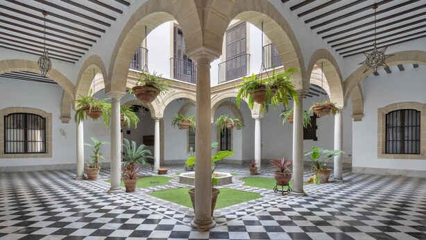 A spacious courtyard with a checkered floor, arches, hanging planters, and a central fountain.