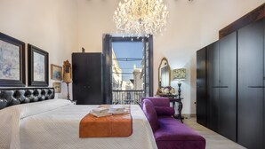 A bedroom with a bed, a purple armchair, a chandelier, and a view of the city through an open door.