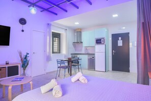 A hotel room with a bed, a small table, a chair, a desk, a TV, a clock, a window, a door, a kitchen area with cabinets, a microwave, and a refrigerator.
