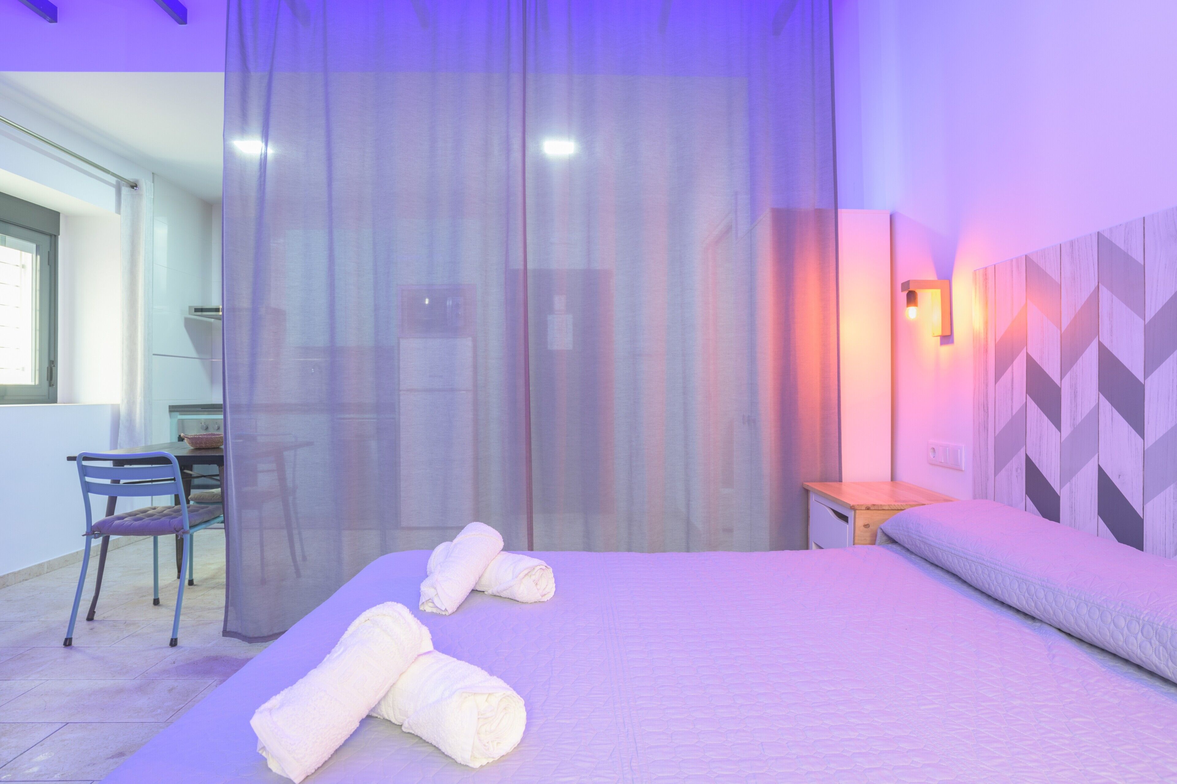 A bedroom with a bed, two rolled towels, a chair, a small table, and a wall-mounted light.