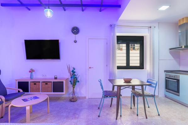 A modern living room with a flat-screen TV, a dining table with chairs, a sofa, a small cabinet, and a kitchen area with a stove.