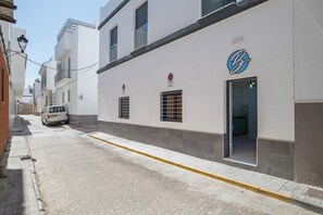 Une rue étroite bordée d’un immeuble blanc orné d’un logo bleu, avec un fourgon garé sur le côté et un trottoir jaune.