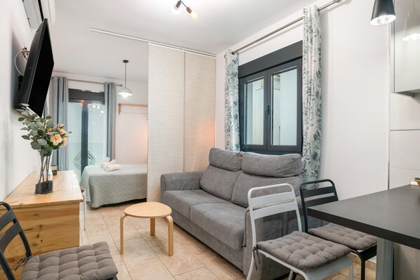 Un salon moderne avec un canapé, une petite table ronde, un téléviseur fixé au mur et une fenêtre avec des rideaux.