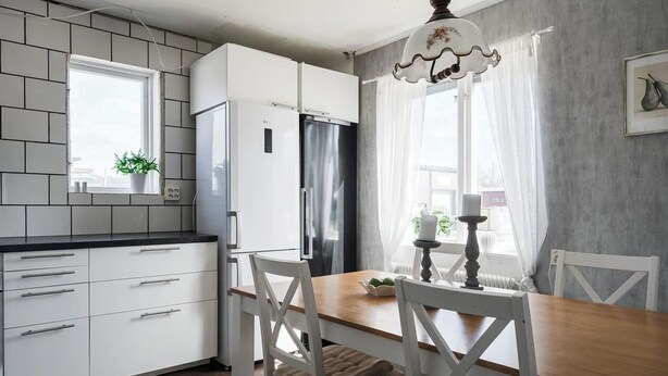 Une cuisine moderne avec des meubles blancs, un réfrigérateur encastré, une table à manger avec des chaises et une fenêtre avec des rideaux.