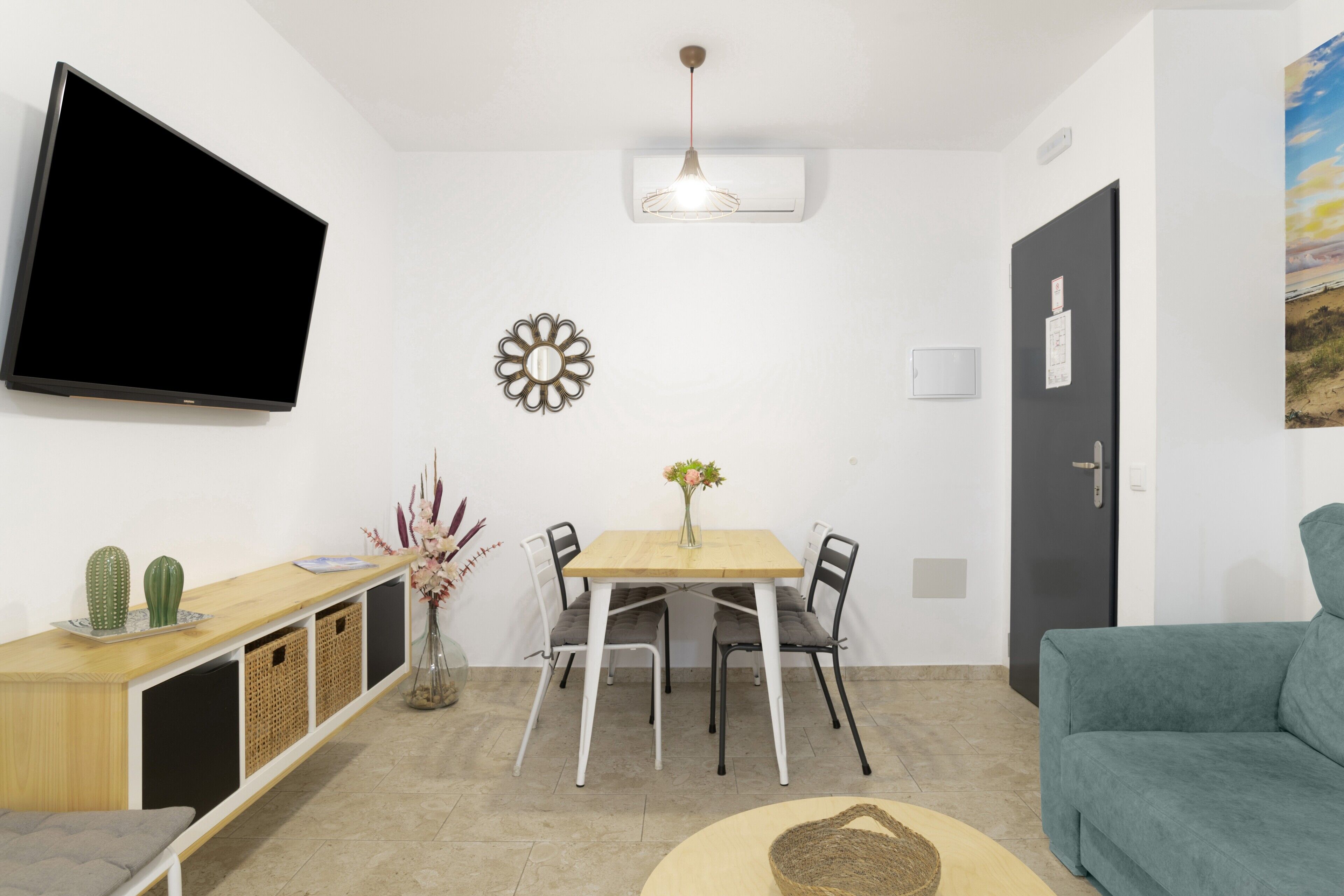 A living room with a flat-screen TV, a dining table with chairs, a sofa, a wall-mounted TV, a ceiling light fixture, and a door.