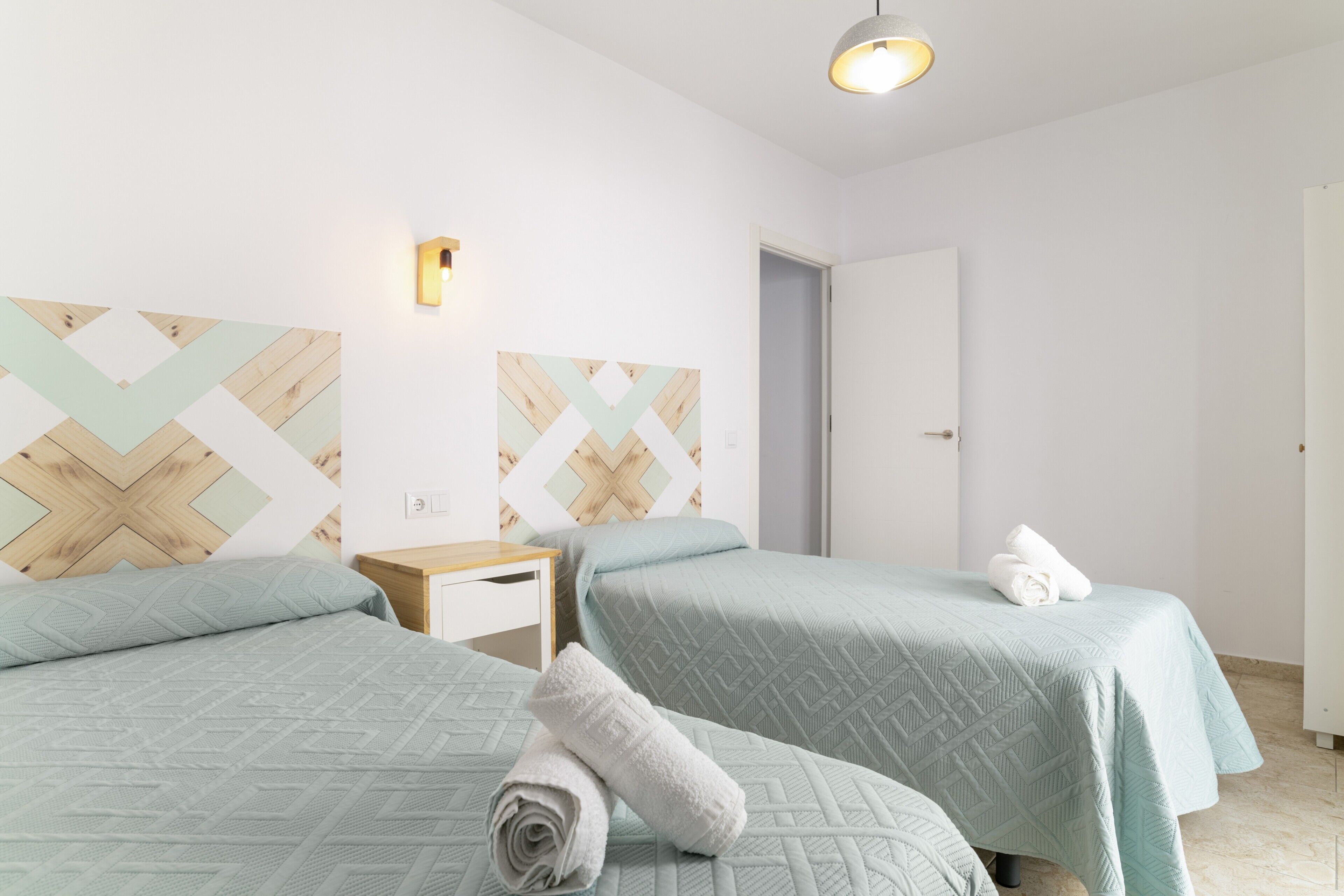 Et hotelværelse med to senge, et natbord og en lampe på væggen.
