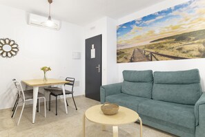 Un salon comprenant un canapé, une table à manger avec des chaises, une petite table ronde avec un panier, un tableau accroché au mur et une porte sur laquelle est apposée une affiche.