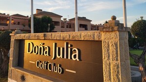 Une enseigne indiquant le club de golf Doña Julia, avec trois drapeaux, un mur de pierre et des bâtiments en arrière-plan.
