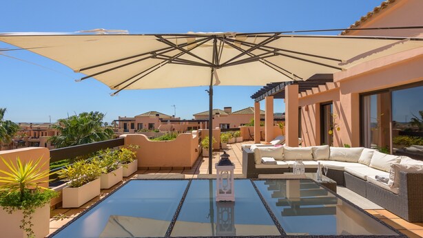 Une terrasse sur le toit avec une table en verre, des meubles en osier et un grand parasol.