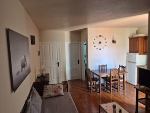 A living room with a sofa, a dining table with chairs, a kitchen area with cabinets and a refrigerator, and a wall clock.