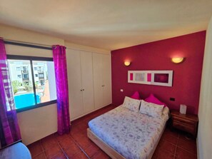 A bedroom with a bed, a window with curtains, a wall-mounted light, and a small bedside table.