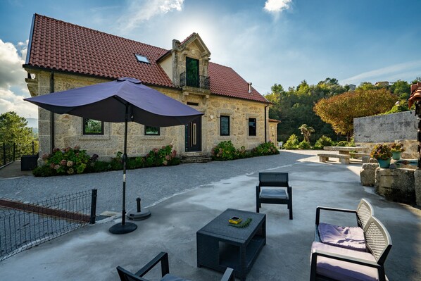 A stone building with a red-tiled roof, outdoor seating with chairs and tables, and a large umbrella.