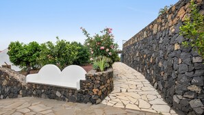 A stone pathway leading to a white bench, surrounded by lush greenery and a stone wall.
