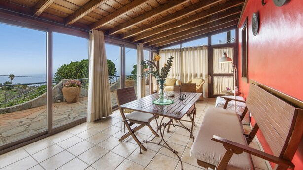 A spacious balcony with a wooden ceiling, a dining table with chairs, a sofa, and a view of the ocean.