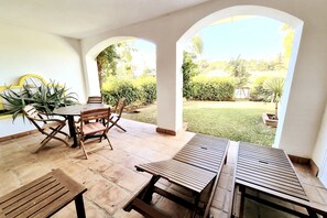 Um alpendre exterior com mobiliário de madeira, incluindo mesa e cadeiras, e vista de um jardim bem cuidado através de um arco.