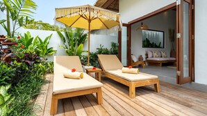A wooden deck with two lounge chairs, a yellow umbrella, and a glass door leading to an interior space with a sofa and a painting. - Villa Lounes 1 (Tabanan)