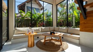 A modern living room with a large window, a sofa, a round wooden table, and a woven rug. - Villa Saya (Canggu)