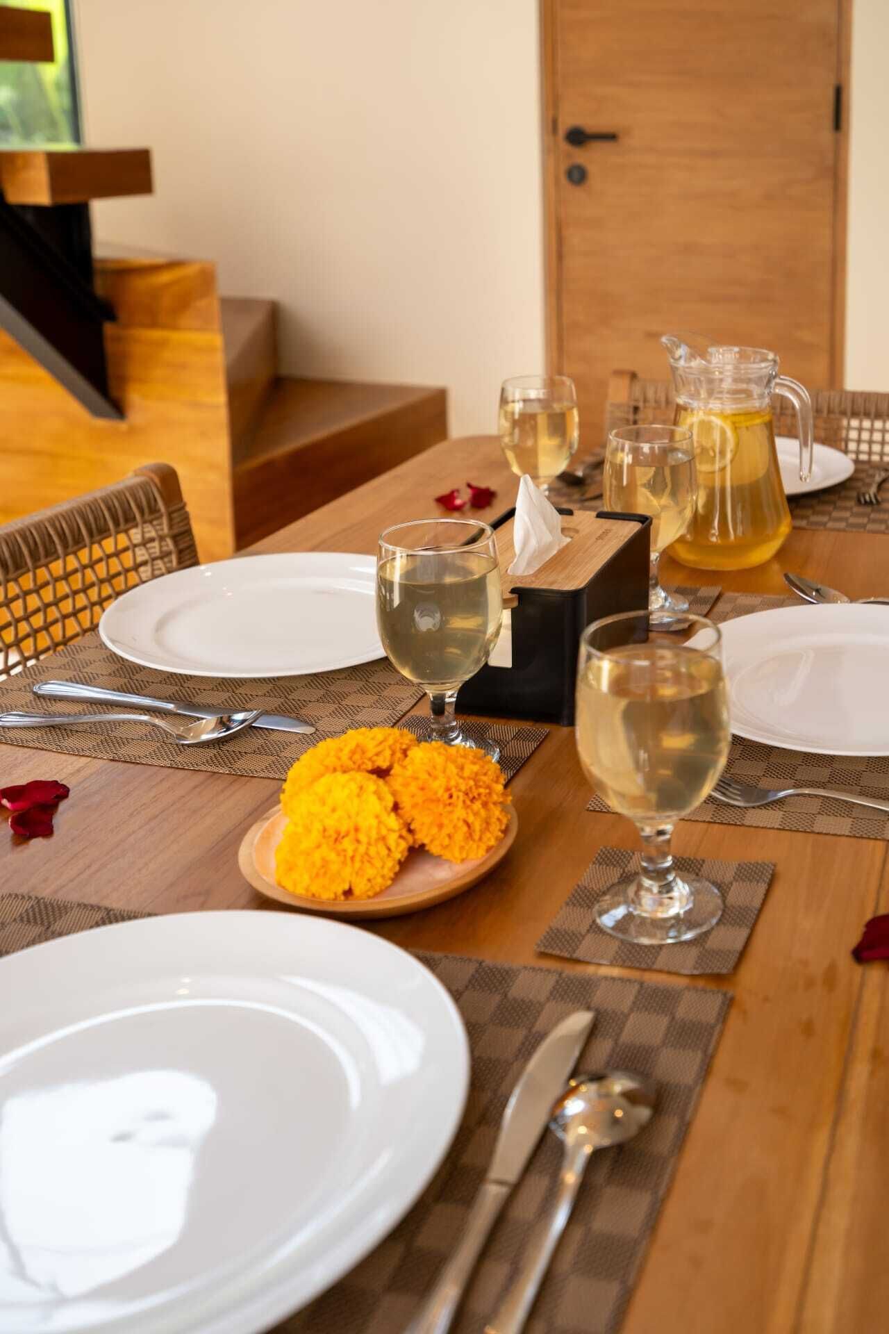 Una mesa de comedor dispuesta con platos blancos, copas de vino y una jarra de bebida amarilla, con un plato de flores amarillas en el centro.