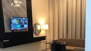 A modern living room with a flat-screen TV mounted on the wall, a round mirror, a floor lamp, a coffee table, and a beige sofa. - Al Zahra Hotel Apartments (Ras Al Khaimah)