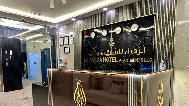 A reception area with a modern design, featuring a reception desk, a sofa, and a wall with clocks and the hotel name. - Al Zahra Hotel Apartments (Ras Al Khaimah)