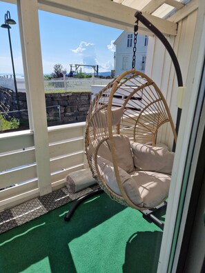 A wicker hanging chair with cushions, a green artificial turf floor, and a view of a white fence and stone wall.