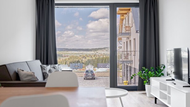 A modern living room with a large sliding glass door leading to a balcony, a sofa, a dining table, and a TV stand. - Plešivec by Interhome (Abertamy)