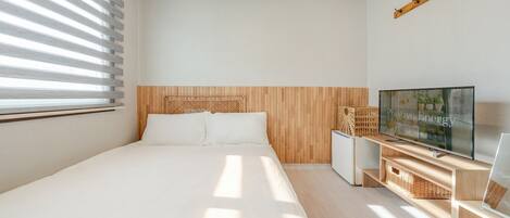 A bedroom with a bed, a TV, a small cabinet, and a window with blinds.