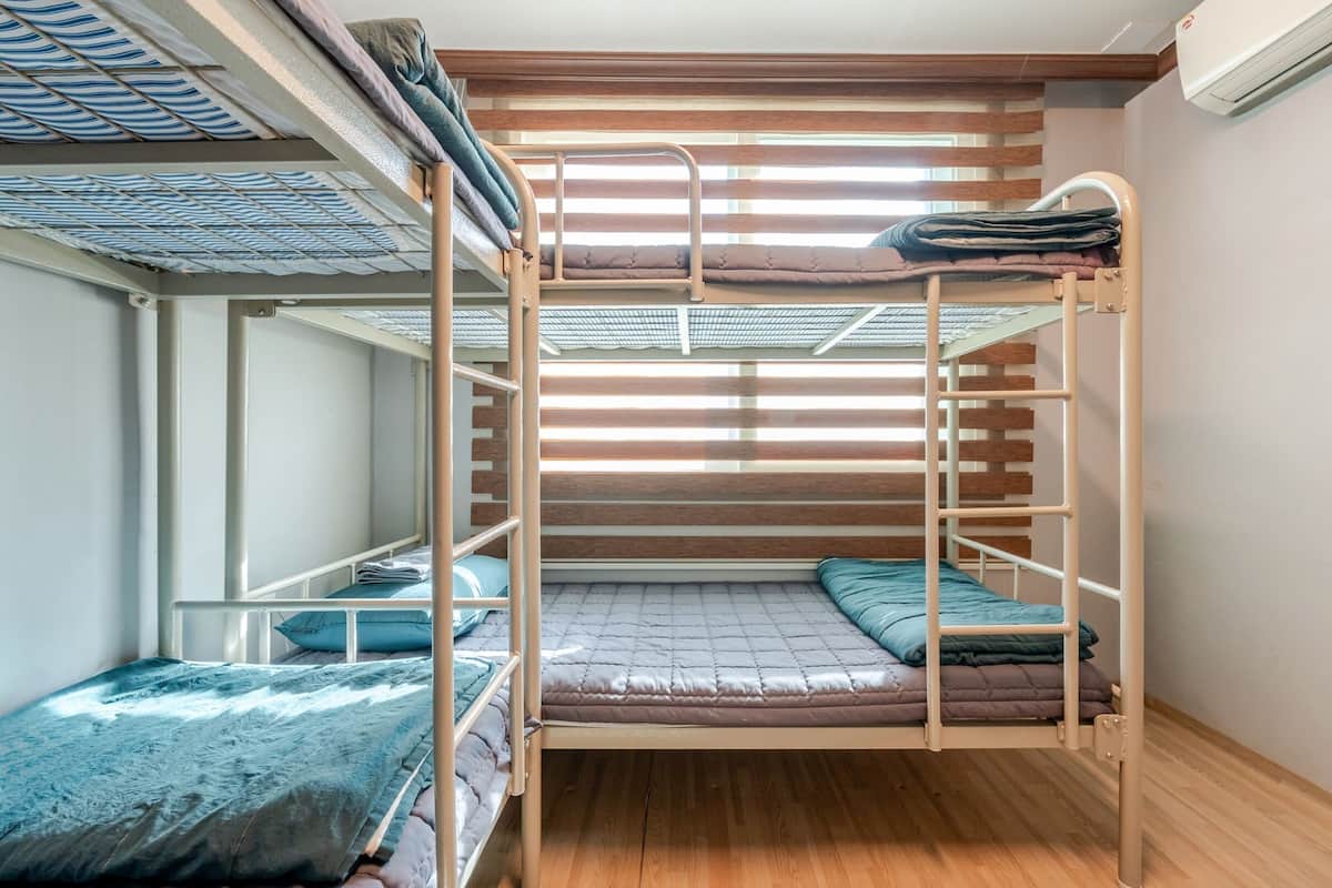 Habitación con literas, piso de madera, barandales de metal y una ventana con persianas.