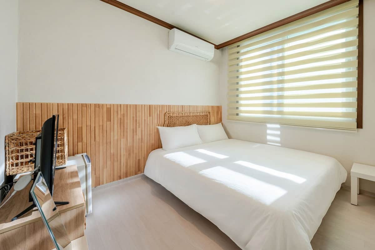 Un dormitorio con cama, televisor, mesita de noche pequeña, lámpara y ventana con persianas.
