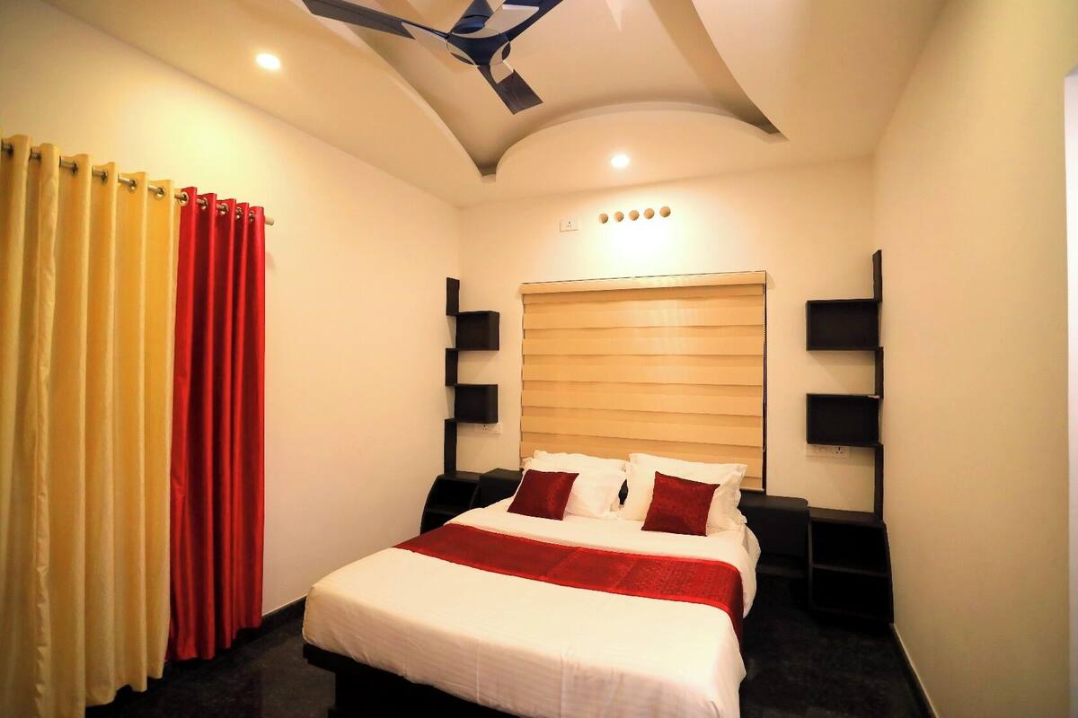 A hotel room with a bed, a ceiling fan, a window with blinds, and a wall-mounted shelf.