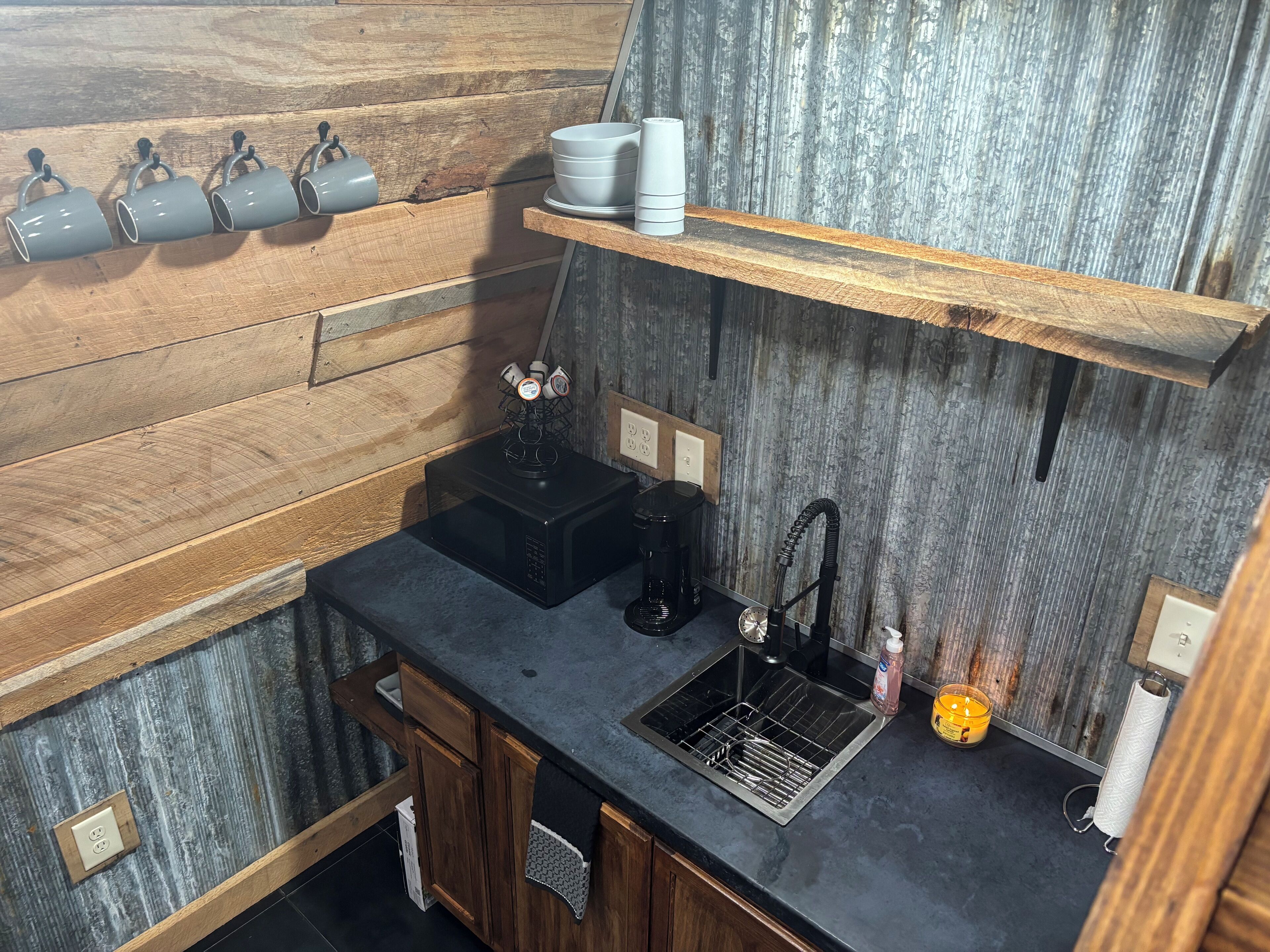 Eine kleine Küche mit schwarzer Arbeitsplatte, Spüle, Kaffeemaschine und Wasserkocher auf einer Holzwandregal.