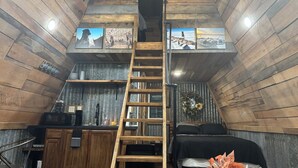 A loft-style bedroom with a wooden ladder leading to a lofted sleeping area, a bed with black bedding, and a small kitchenette with a microwave and sink. - Peaceful secluded cabin located near Horseshoe Lake  (Miller City)