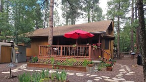 A wooden cabin with a deck, surrounded by a garden with raised beds and a stone pathway. - Garden Cabin with Fenced Yard and AC (Pinetop)