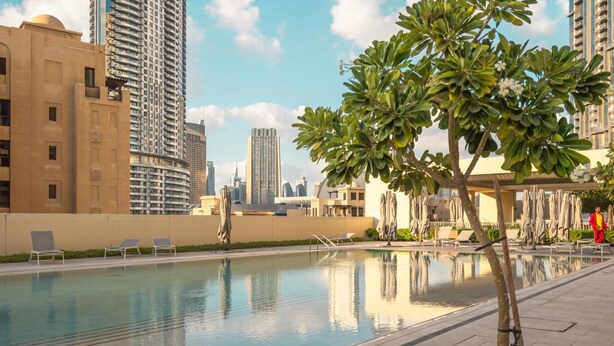 A swimming pool with lounge chairs and umbrellas, surrounded by tall buildings and a clear sky.