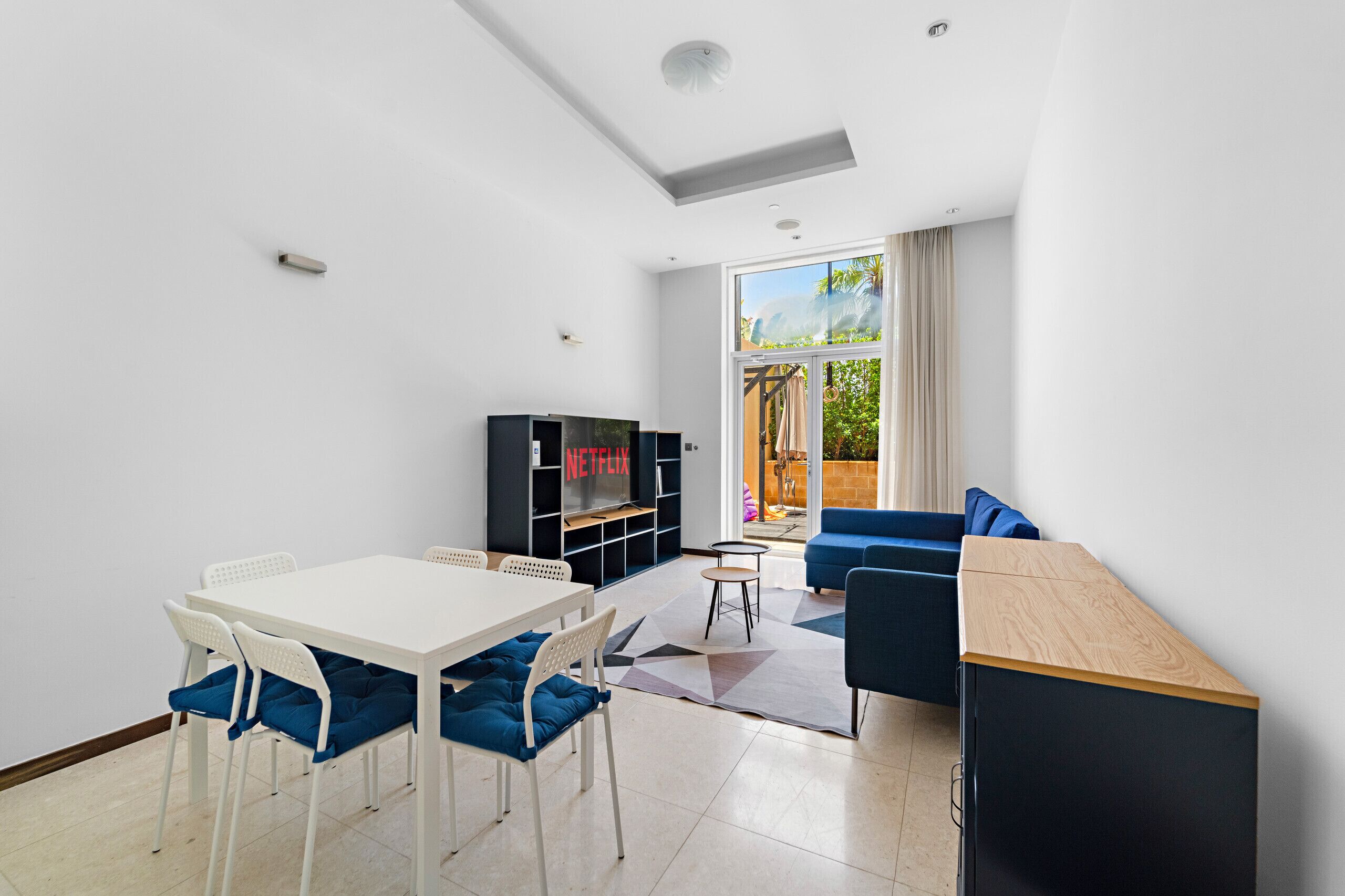 A modern living room with a white table, blue chairs, a blue sofa, a black entertainment unit, and a large window with a view of greenery.