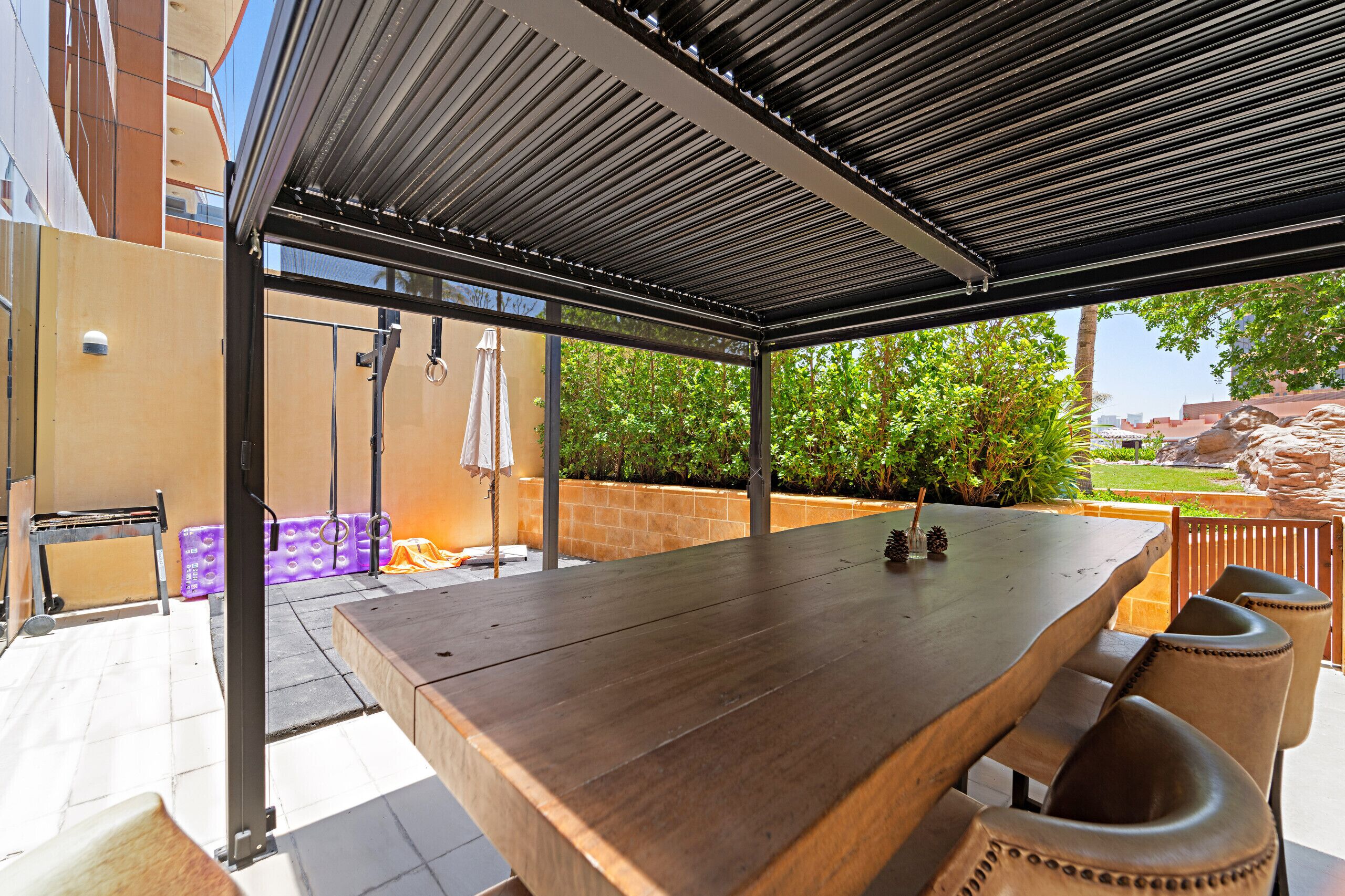 Un patio techado al aire libre con una larga mesa de madera y sillas, un banco y un pequeño jardín con plantas y vistas a la ciudad.