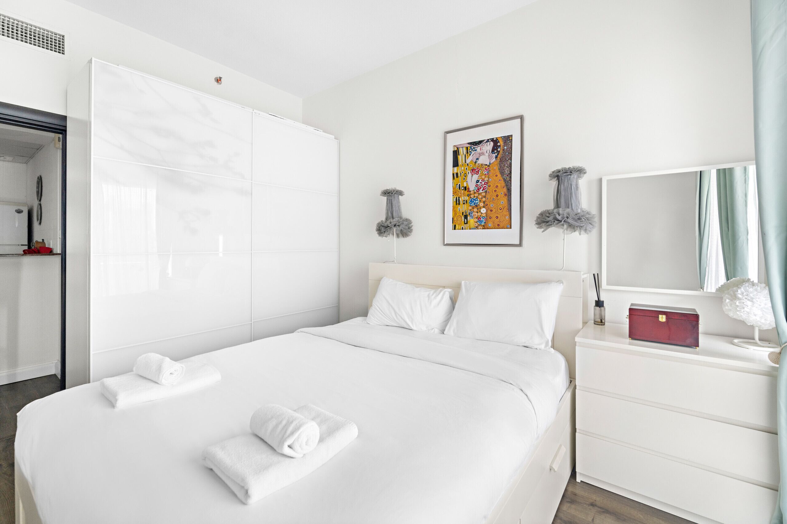 Une chambre d’hôtel avec un grand lit, des murs blancs, un tableau encadré, une commode et un miroir.