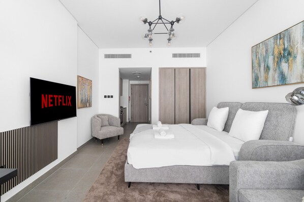 A modern bedroom with a large bed, a grey sofa, a TV displaying 'NETFLIX', and a painting on the wall. - Modern Studio with Great Amenities (Dubai)
