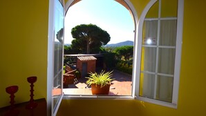 A yellow room with a large arched window overlooking a garden with a tree and a brick structure. - Villa Petit Castell 10 people Costa Brava (PALAFOLLS)