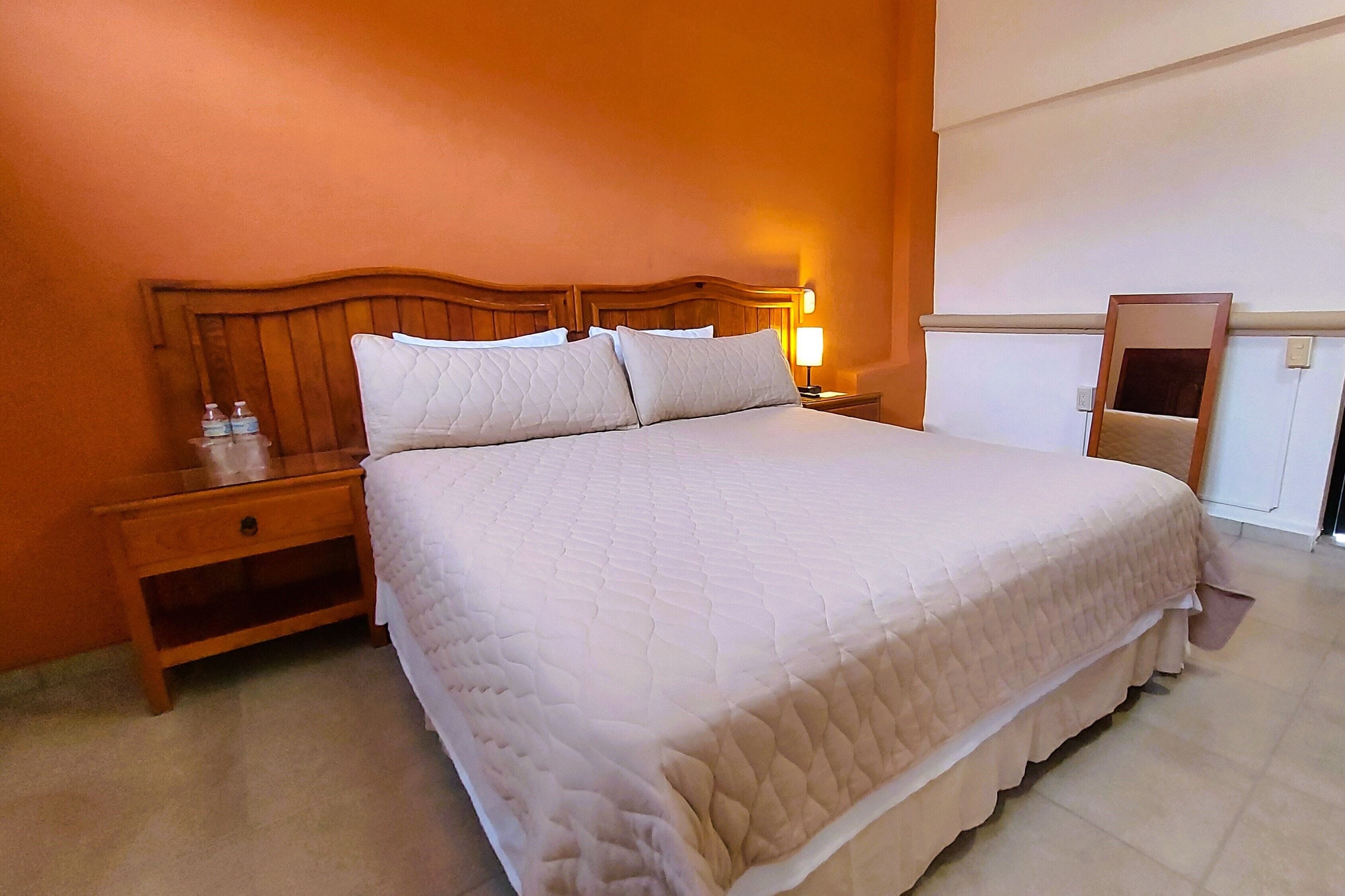 Een netjes opgemaakt bed met een houten hoofdbord, een nachtkastje met een lamp en een spiegel aan de muur.