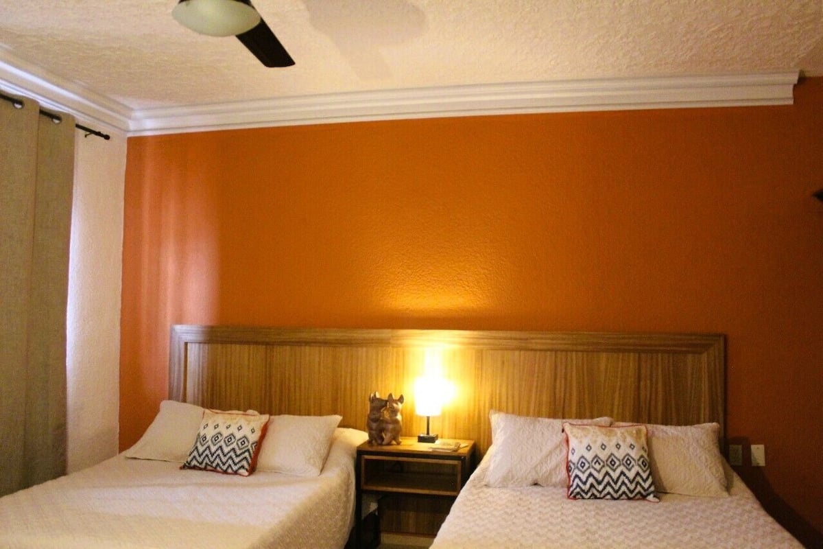 Quarto de hotel com duas camas, uma mesa de cabeceira com abajur e um ventilador de teto.
