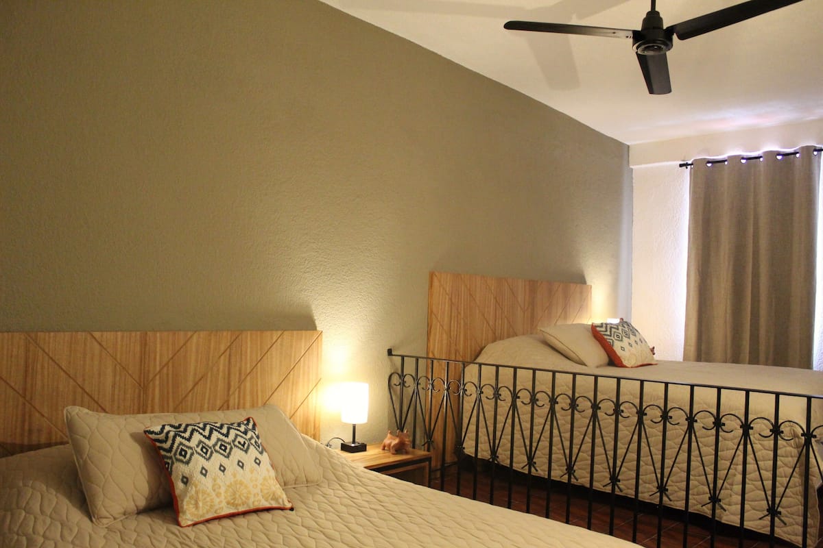 Quarto de hotel com duas camas, um ventilador de teto, mesa de cabeceira com abajur e janela com cortinas.