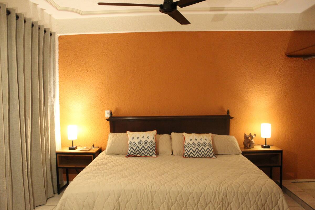 Quarto de hotel com uma cama grande, duas mesas de cabeceira com abajures, um ventilador de teto e vista para as paredes laranjas do quarto.