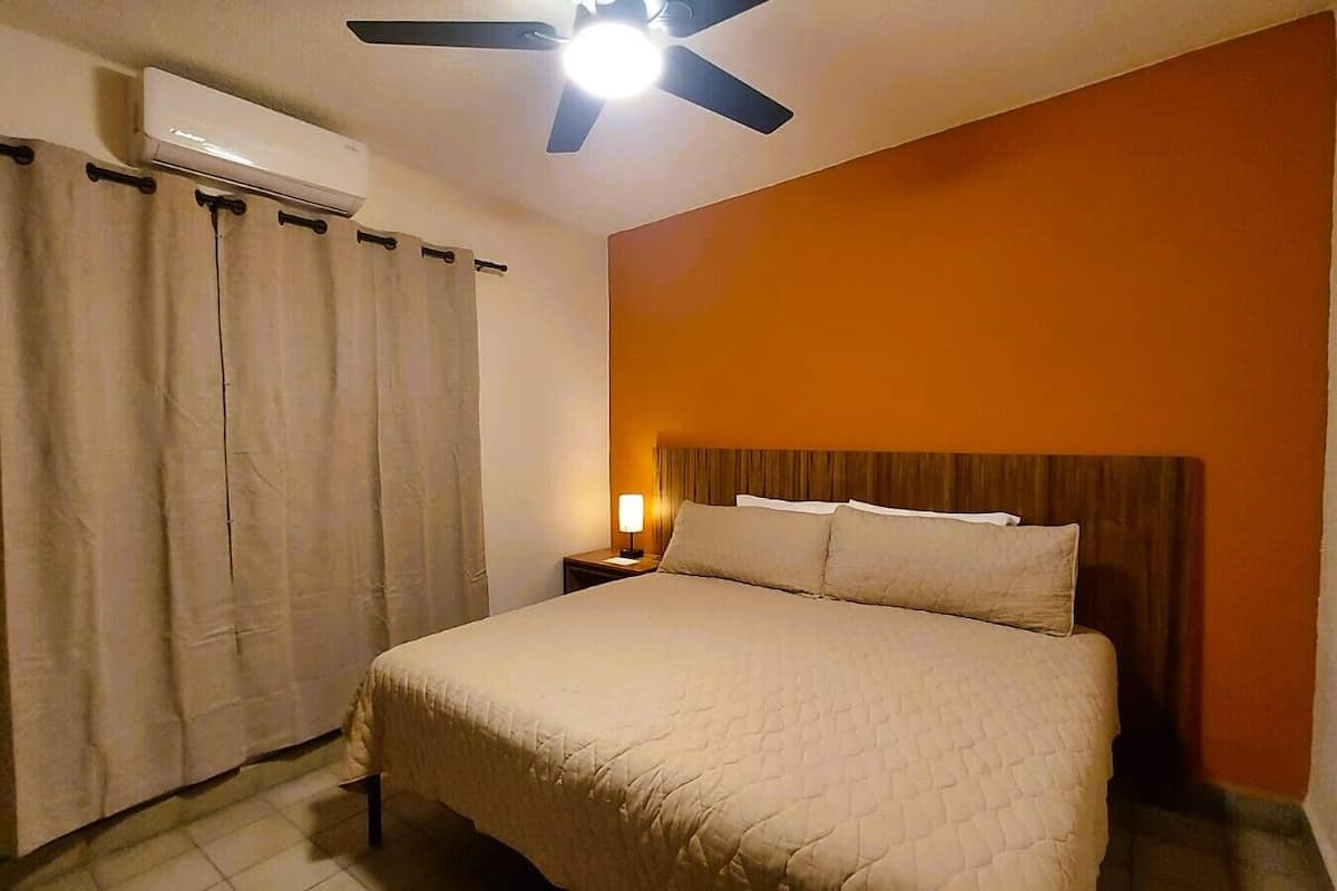 Quarto de hotel com cama, ar condicionado, mesa de cabeceira com luminária e ventilador de teto.