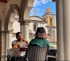 Dos personas sentadas en un café al aire libre, con vista a un edificio histórico de cúpula amarilla y una estatua.