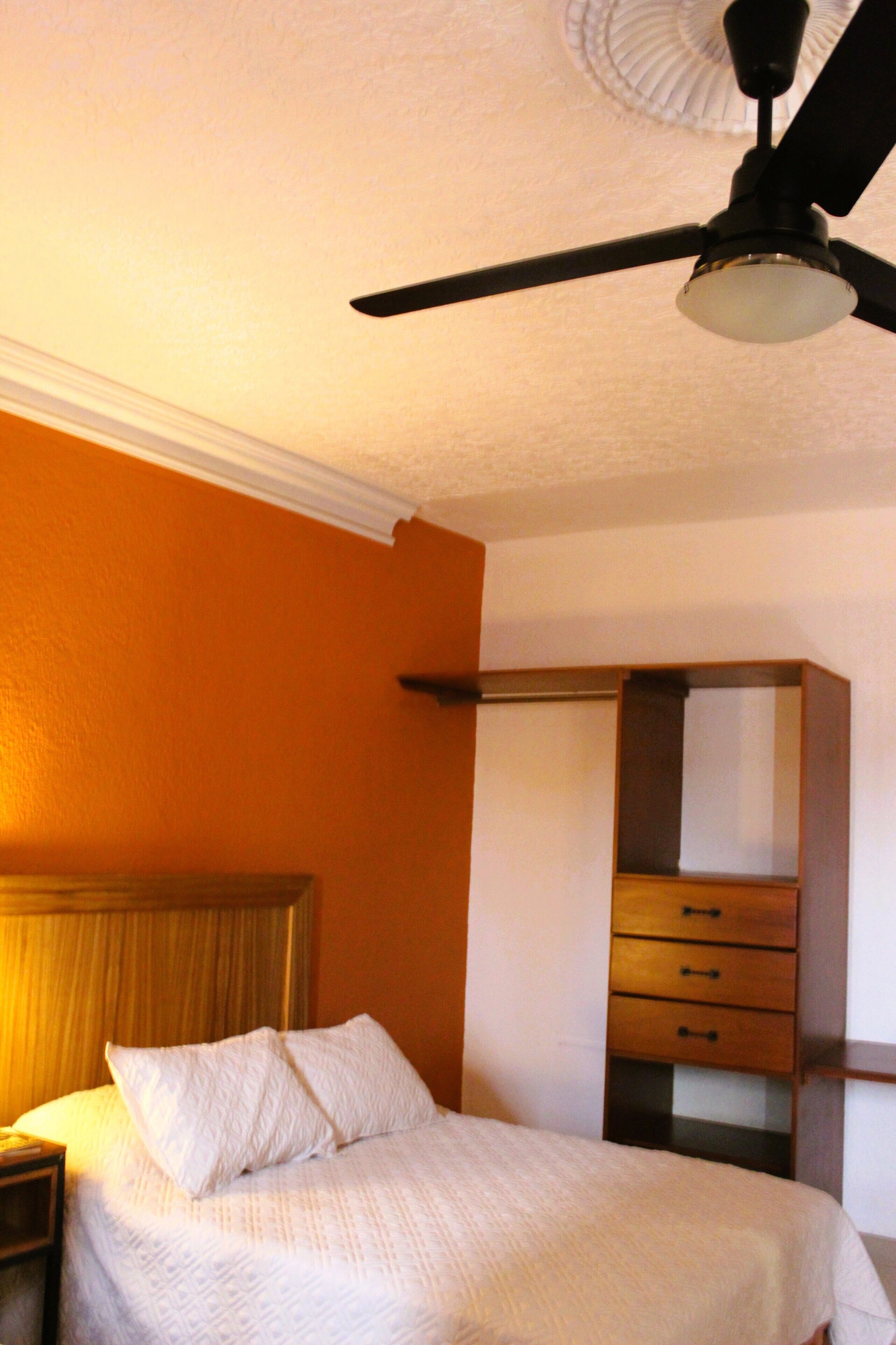 Un dormitori amb llit, una tauleta de nit, una commode i un ventilador de sostre.