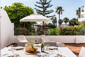 Un espace de restauration en plein air avec une table dressée pour deux personnes, une bouteille de vin et un parasol blanc.