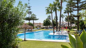 A swimming pool surrounded by palm trees and lounge chairs, with a thatched-roof structure nearby.